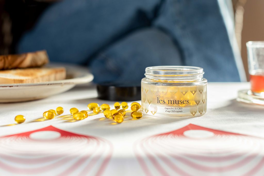Quand on se lève, on peut prendre du CBD pendant son petit déjeuner : un pot de gélules dorées est posé sur une table avec une nappe blanche avec des tartines et du miel.