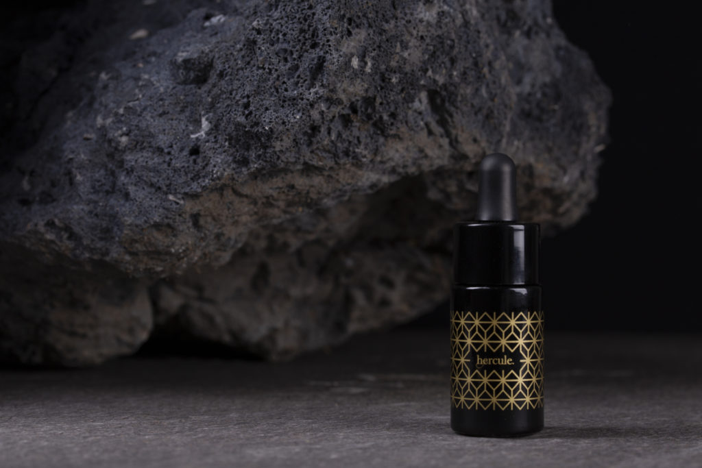 Un flacon noir sérigraphié de motifs dorés avec au centre, le nom du produit, hercule. Le flacon est posé sur un fond en pierre avec en arrière-plan, une roche volcanique.